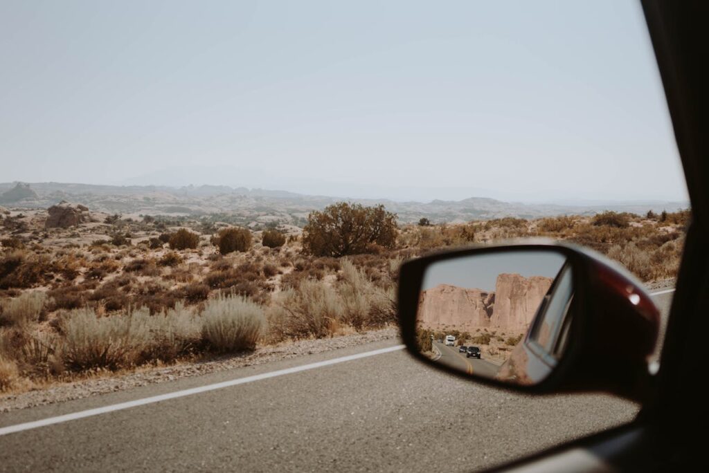 View from a car mirror looking back at a desert highway surrounded by rocky terrain during a long road trip through nature.