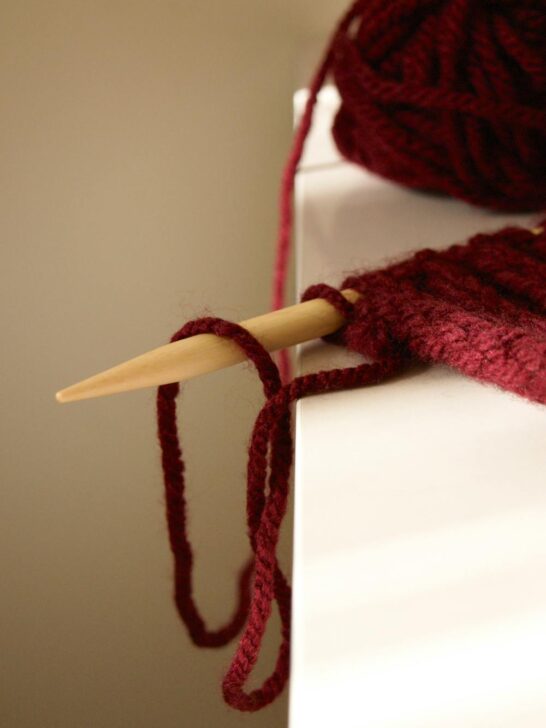 Close-up of knitting project with red yarn and wooden needle resting on table edge