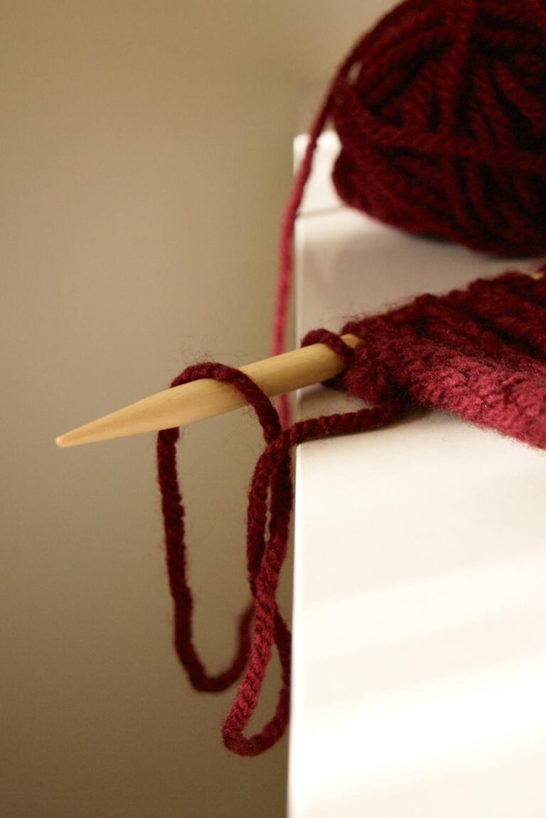 Close-up of knitting project with red yarn and wooden needle resting on table edge