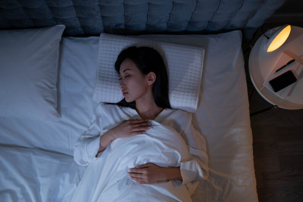 Woman peacefully sleeping on her back in a cozy bed with soft pillows and warm bedside lighting