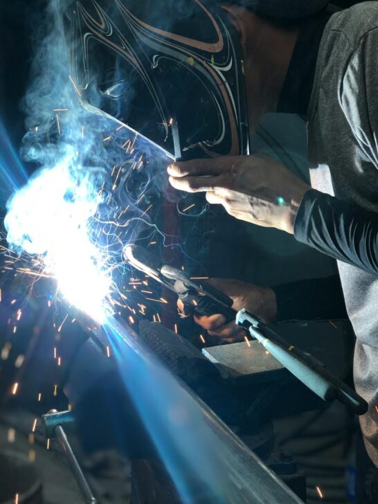 Close-up of welder using torch on metal pipe with bright sparks and protective helmet in a dim workspace