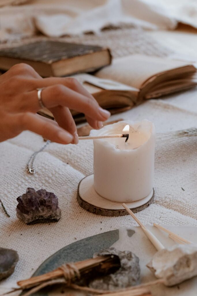 Hand lighting a white candle on a cozy table with crystals, books, and soft neutral decor creating a calm atmosphere