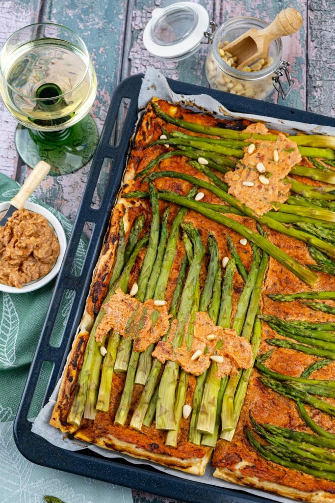 Baked flatbread topped with asparagus and sauce on a sheet pan with a glass of white wine nearby