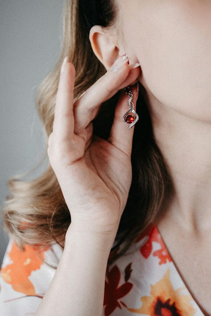 Woman wearing silver charm earrings with red gemstone while adjusting jewelry near her ear