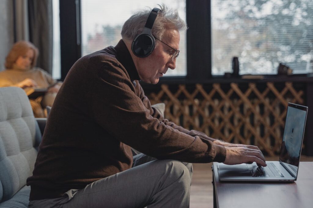 Older man using a laptop with headphones while sitting in a comfortable living space