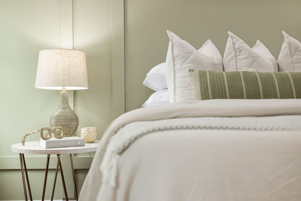 Soft sage green bedroom featuring white bedding, layered pillows, a decorative accent pillow, and a bedside table with a lamp for a calm and relaxing look.