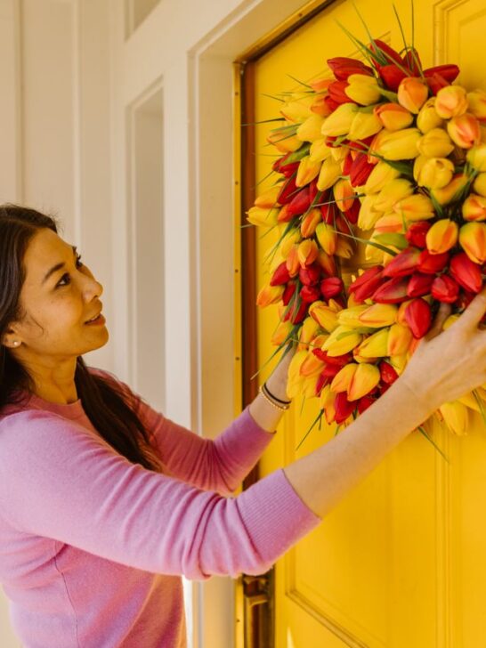 Woman hanging a colorful tulip wreath on a bright yellow front door as part of a simple spring home decor refresh.