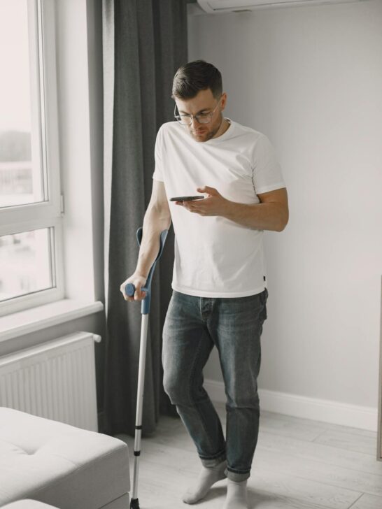 Person using a crutch while checking a smartphone during recovery from delayed injury symptoms after a car accident.
