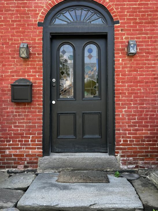 Black arched front door with decorative glass panels set in a historic red brick exterior wall
