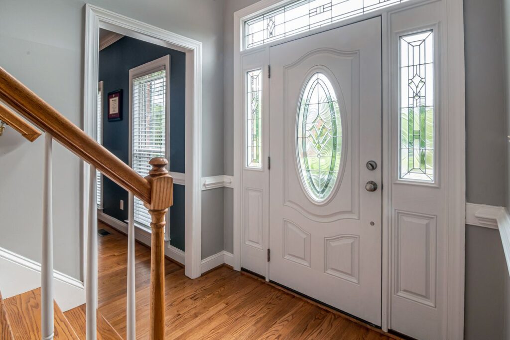 White front door with decorative oval glass insert, sidelights, and transom windows in a bright entryway with hardwood floors