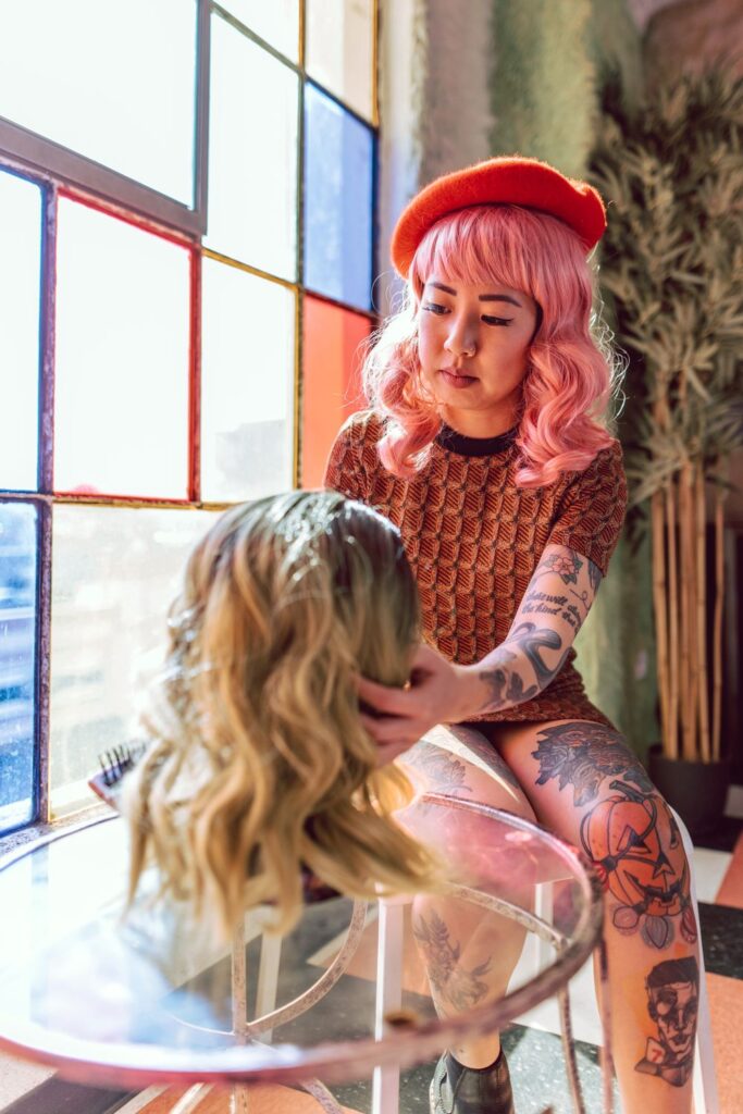 Person holding a blonde wig while wearing a pink wig and orange beret in a brightly lit room with colorful windows.