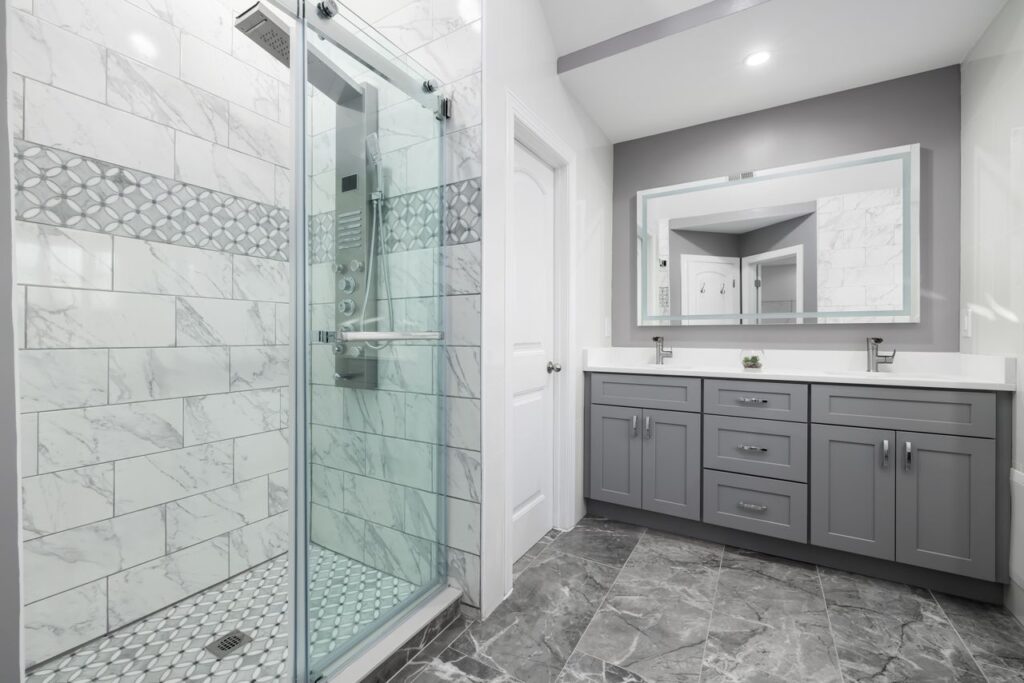 Modern bathroom featuring smart shower technology, marble-look tile walls, glass shower enclosure, and gray double vanity.