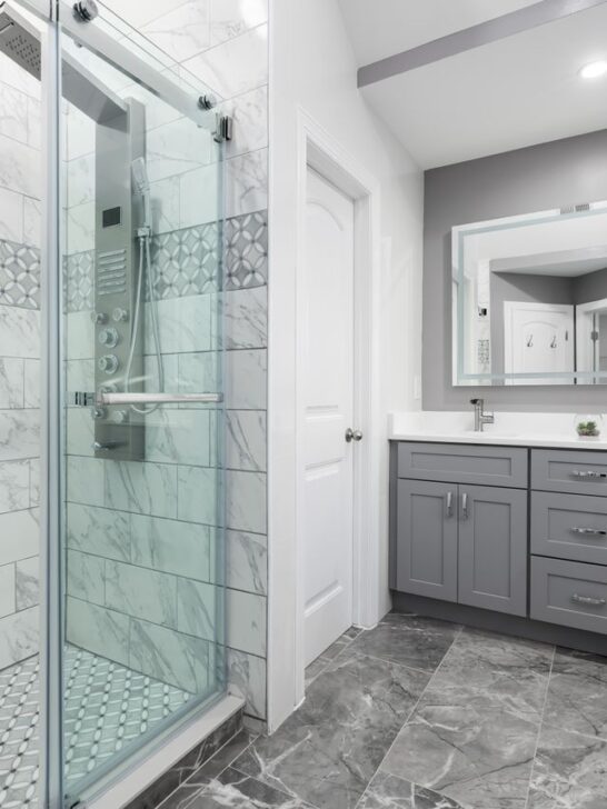 Modern bathroom featuring smart shower technology, marble-look tile walls, glass shower enclosure, and gray double vanity.