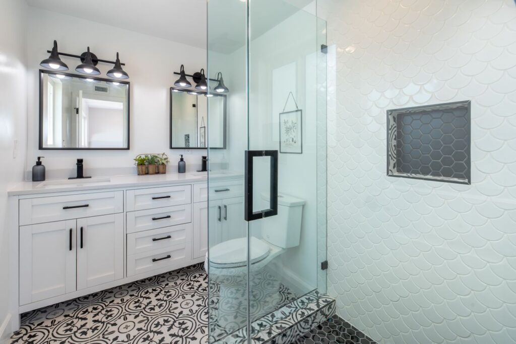 Bright white bathroom with patterned tile flooring, double vanity, glass shower, and decorative shower niche.