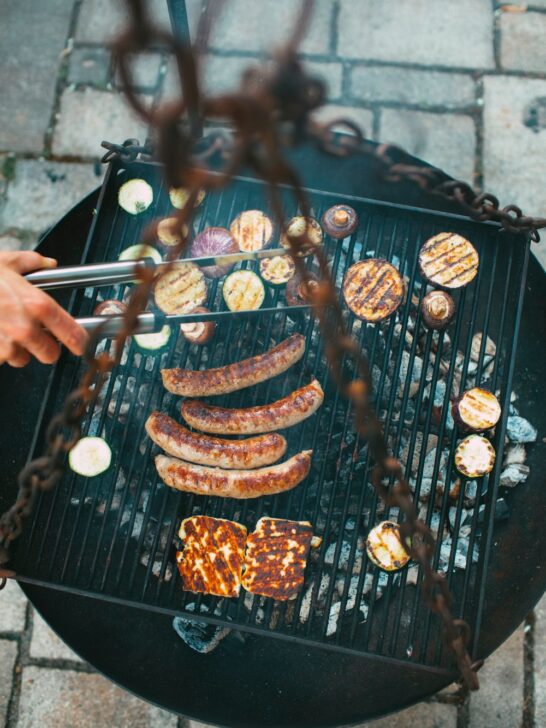 Charcoal grill cooking sausages, grilled vegetables, and cheese over hot coals during an outdoor cookout.