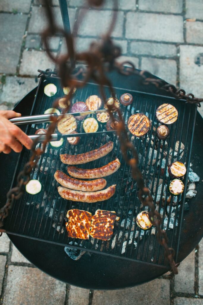 Charcoal grill cooking sausages, grilled vegetables, and cheese over hot coals during an outdoor cookout.
