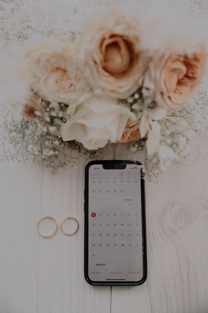 Wedding rings and a smartphone displaying a calendar beside a bridal bouquet for wedding planning.