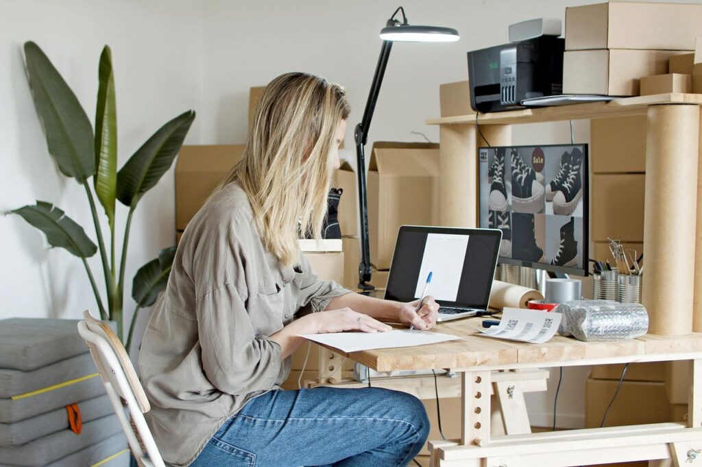 Home business owner organizing orders and paperwork while working from a home office with shipping supplies and a laptop.