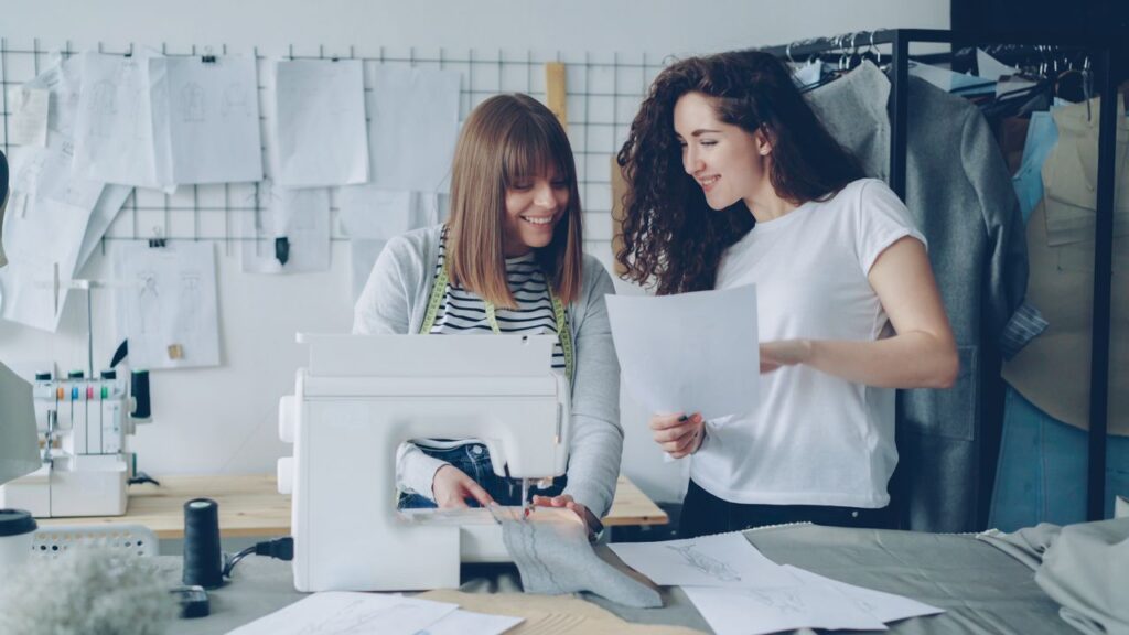 Small business growth meeting with two business owners reviewing paperwork beside a sewing machine in a creative workspace.