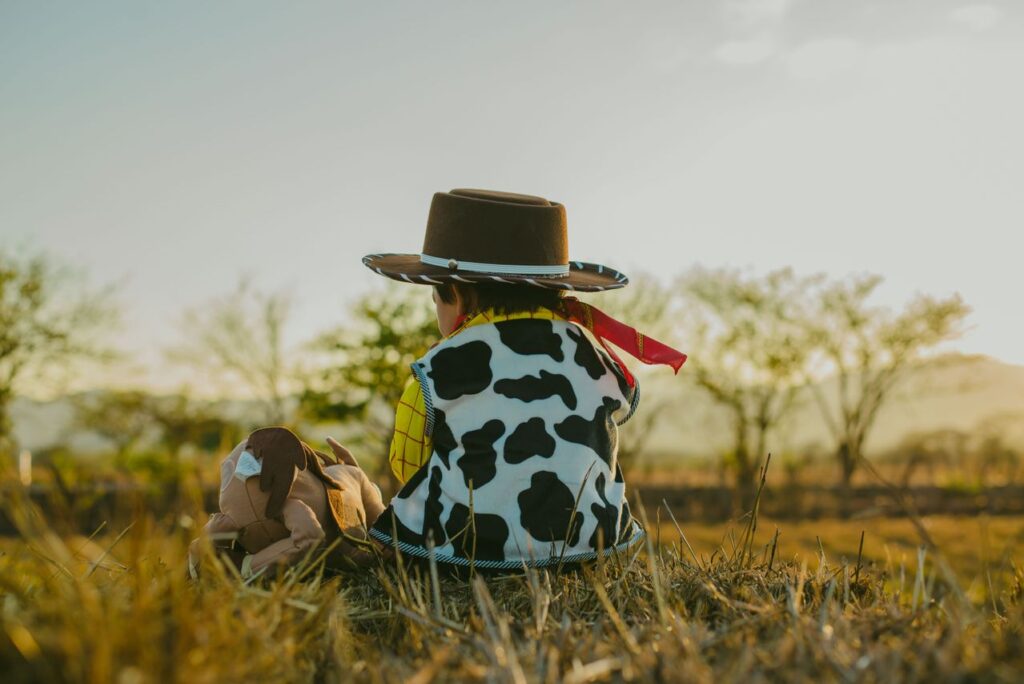 Child dressed as Woody from Toy Story sitting outdoors with a Woody doll in a grassy field
