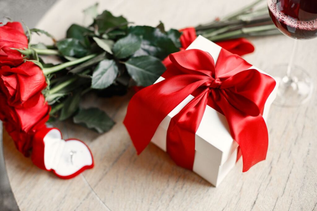 Anniversary gift baskets with red roses, a white gift box wrapped in a red ribbon, and a glass of red wine for a romantic celebration