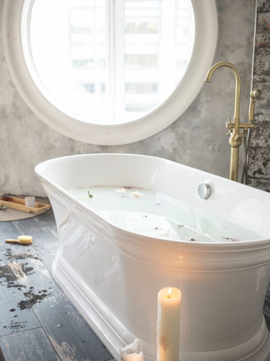 White freestanding soaking tub with brass fixtures, candles, and a large round bathroom window.