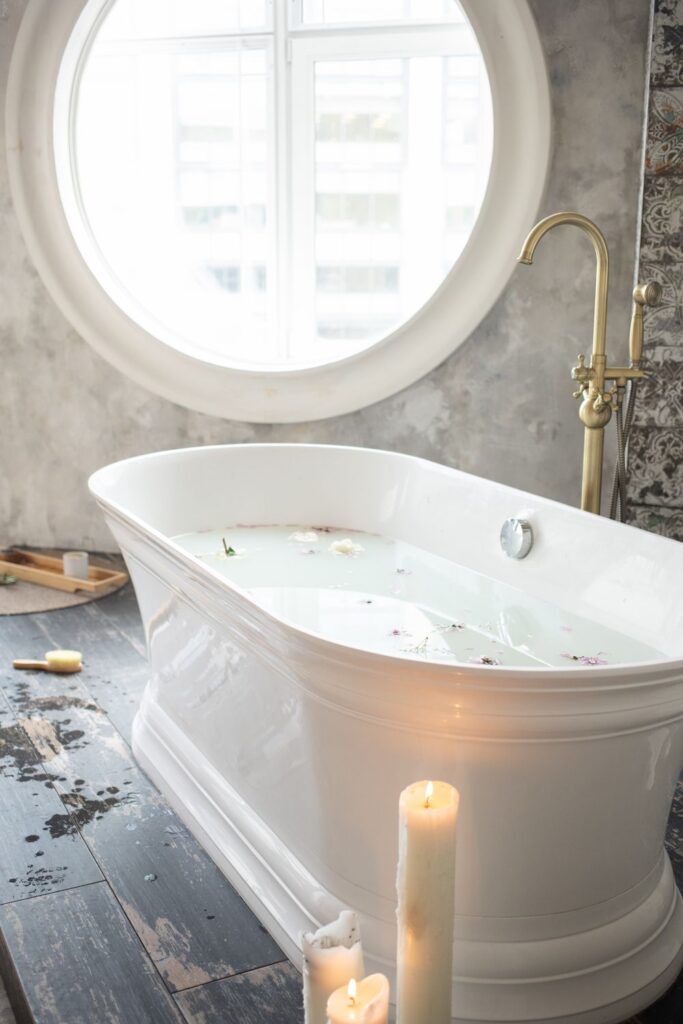 White freestanding soaking tub with brass fixtures, candles, and a large round bathroom window.