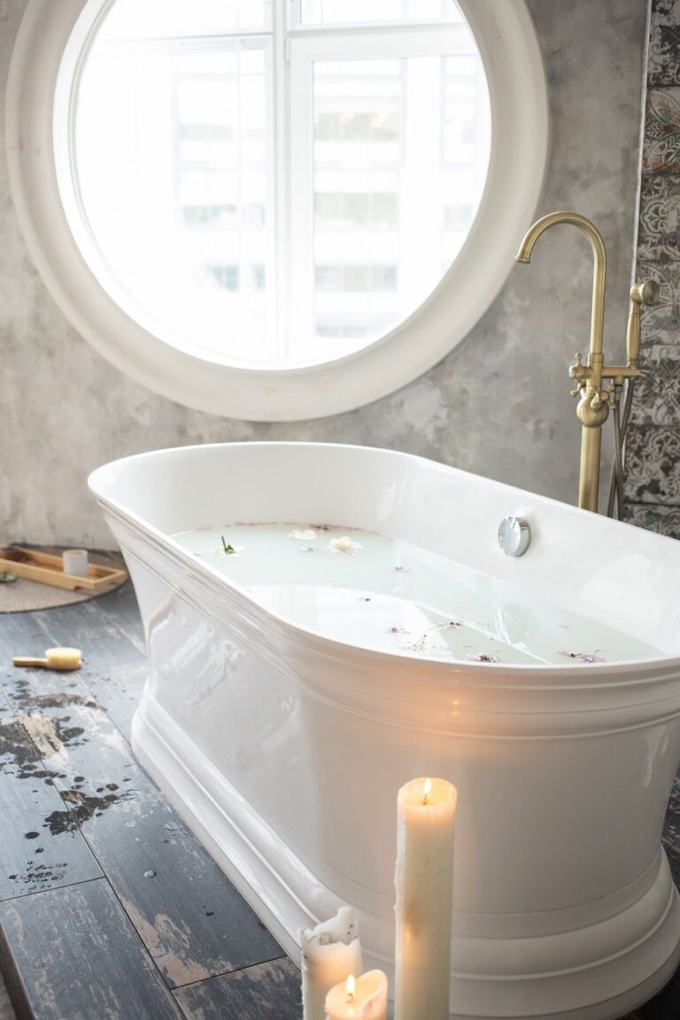 White freestanding soaking tub with brass fixtures, candles, and a large round bathroom window.
