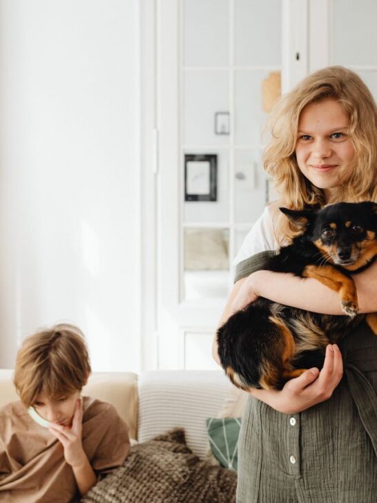 A young girl hugs her small black and brown dog while a boy sits nearby at home.