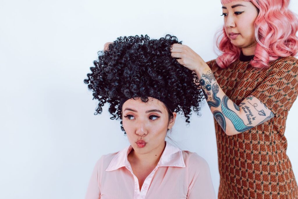 Stylist adjusting a voluminous curly black wig on a woman during a hair styling session.