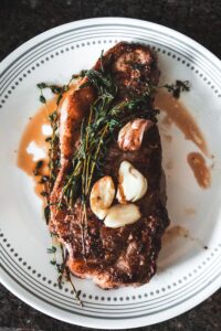 Grilled steak topped with roasted garlic and fresh herbs showing how to order steak based on preferred cut and preparation.