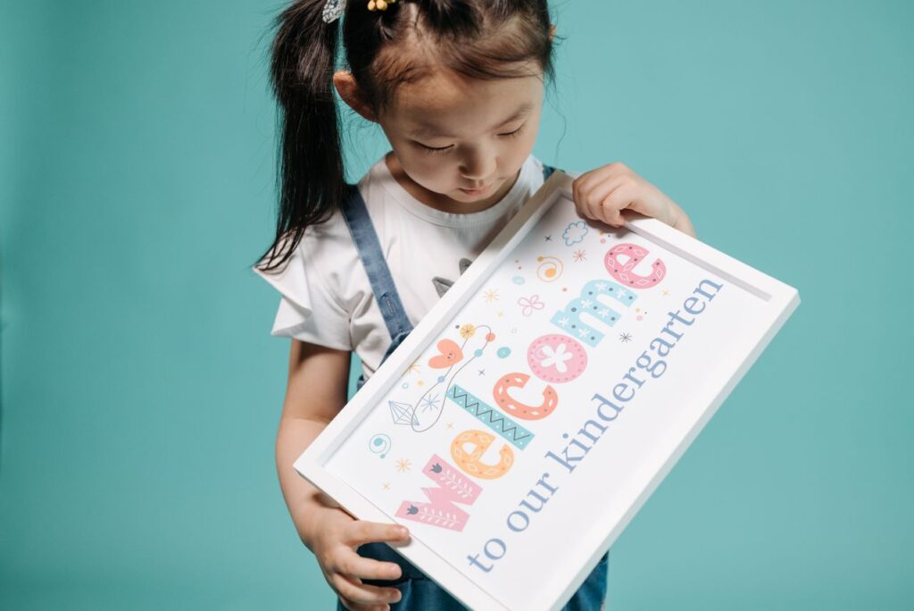 Young student holding a welcome to kindergarten in Singapore sign against a blue background.