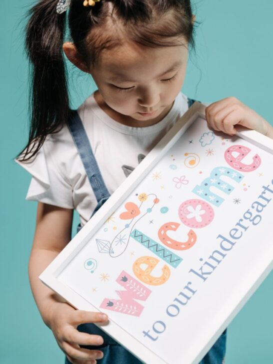 Young student holding a welcome to kindergarten in Singapore sign against a blue background.