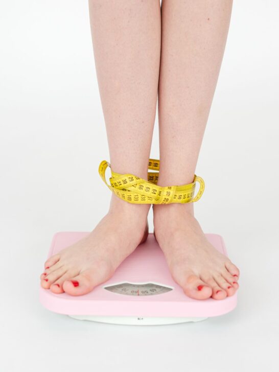 Woman standing on a scale with a measuring tape around her ankles during a weight management journey.
