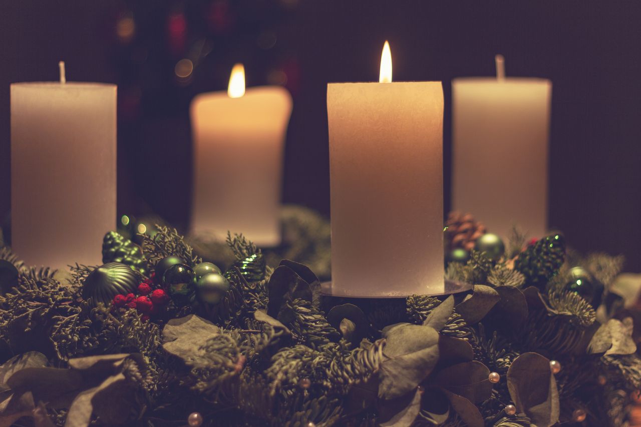 Four ivory pillar candles surrounded by evergreen branches, green ornaments, pinecones, and red berries.