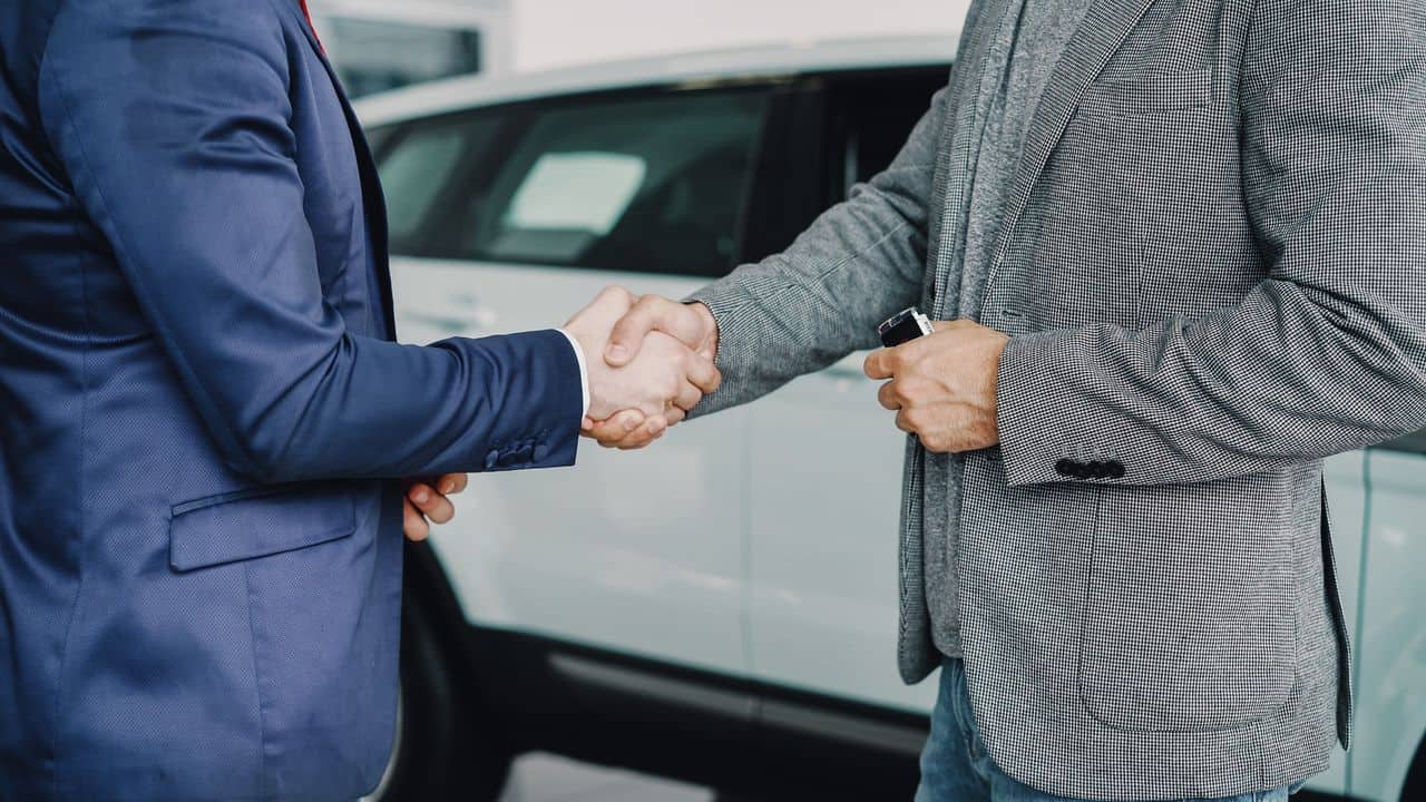 Two people shaking hands beside a white car, with one holding a key fob.