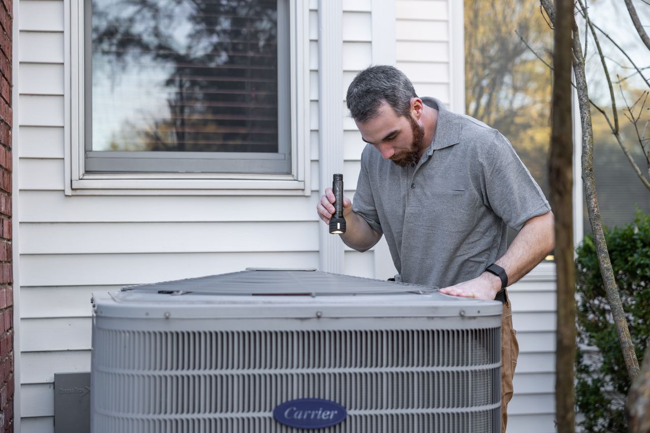 Man inspecting an outdoor unit with a flashlight during heat pump maintenance.