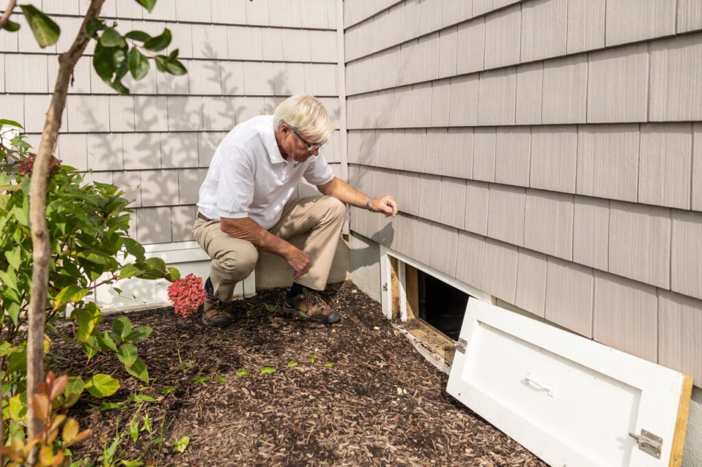 Man inspecting gray house siding beside an open crawl space access door.