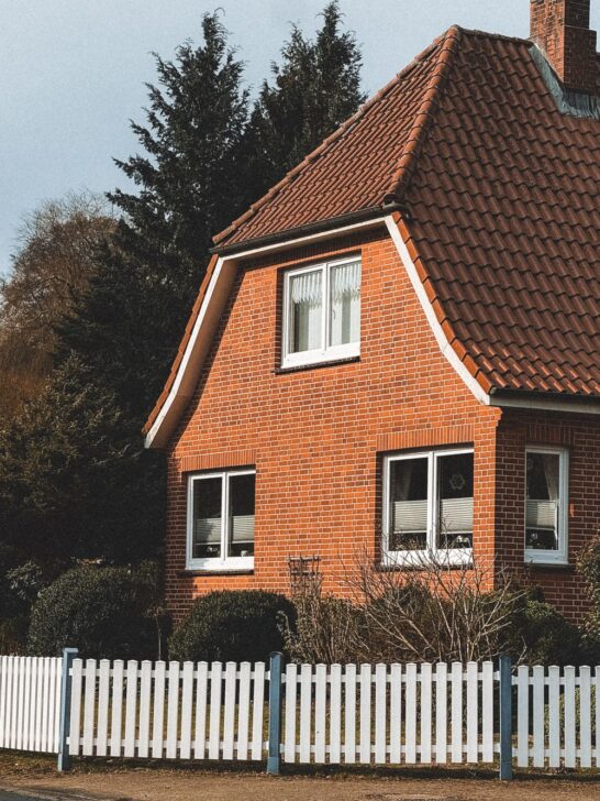 Red brick house with a tiled roof, brick chimney, white-framed windows, and a white picket fence.