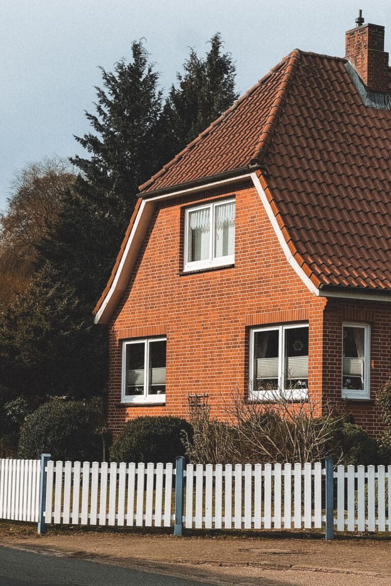 Red brick house with a tiled roof, brick chimney, white-framed windows, and a white picket fence.