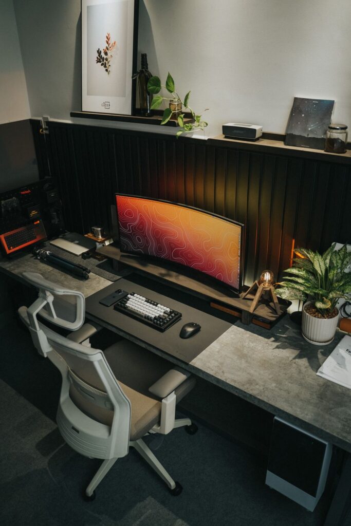 Desk with a curved monitor, keyboard, mesh office chair, potted plants, and a small accent lamp.