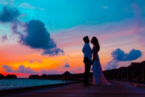 Couple holding hands on a waterfront boardwalk beneath a colorful sunset sky.