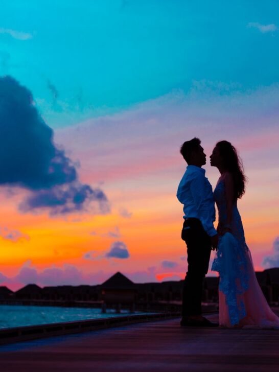 Couple holding hands on a waterfront boardwalk beneath a colorful sunset sky.