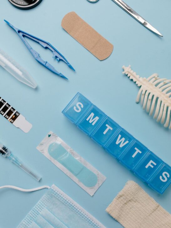 Weekly pill organizer, bandages, medical scissors, face mask, and other medical supplies on a light blue background.