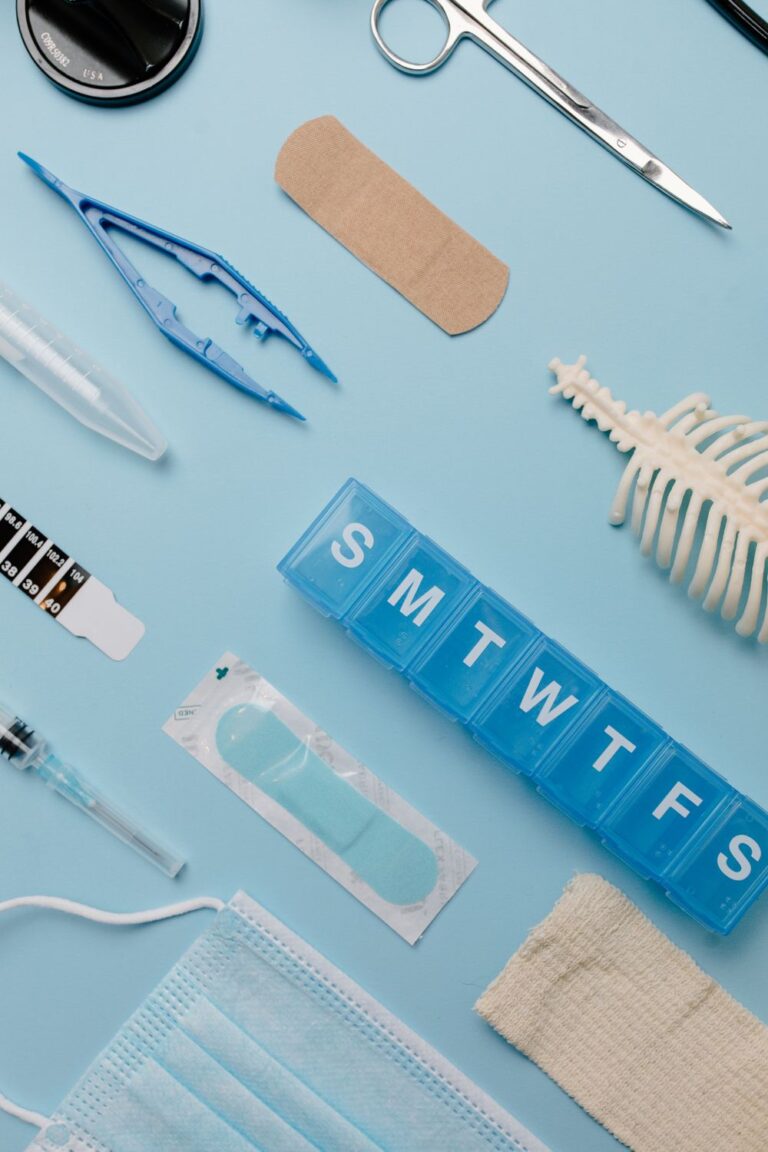 Weekly pill organizer, bandages, medical scissors, face mask, and other medical supplies on a light blue background.