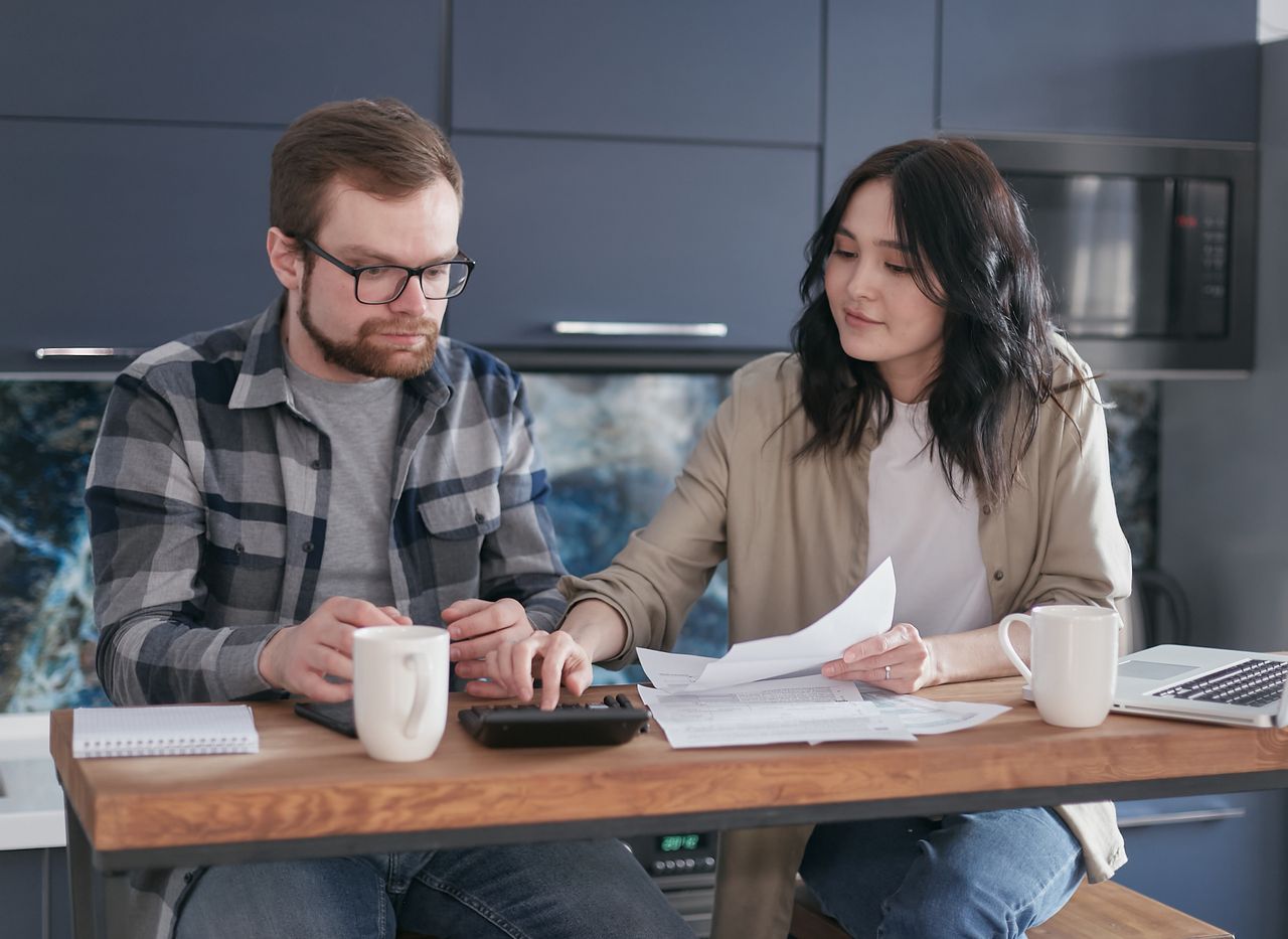 Couple planning family expenses at a kitchen table with bills, a calculator, and a laptop.