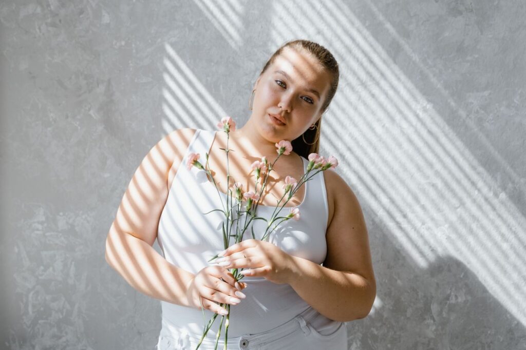 Woman in a white tank top and light jeans holding pink flowers in striped sunlight.