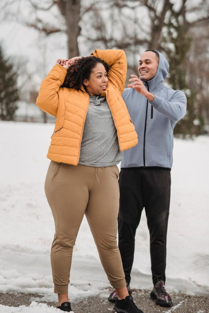 Woman in an orange puffer jacket and tan joggers stretching beside a man on a snowy outdoor path.