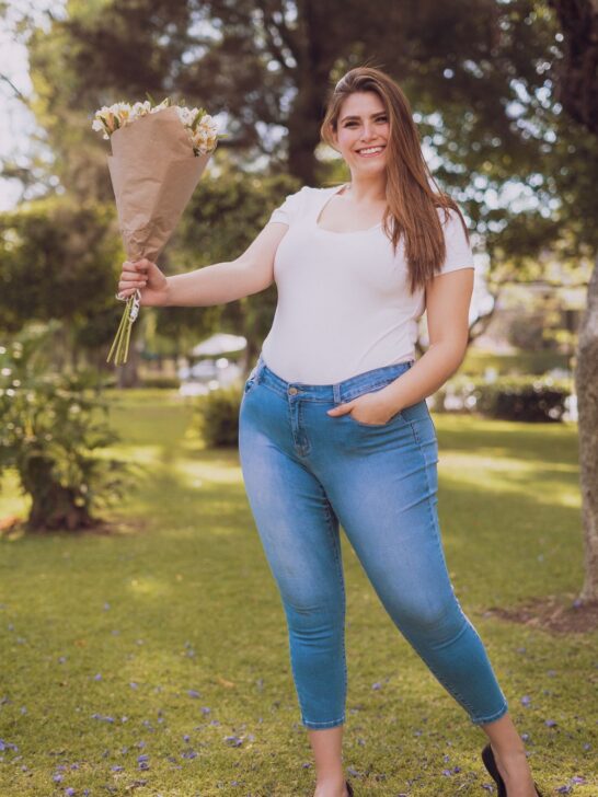 Woman wearing plus-size wardrobe staples&mdash;a white T-shirt, blue jeans, and black flats&mdash;holding a bouquet outdoors.
