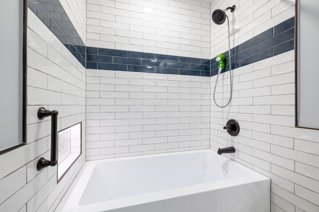 Bathtub and shower with white subway tile, a navy accent stripe, dark fixtures, a grab bar, and recessed storage niche.
